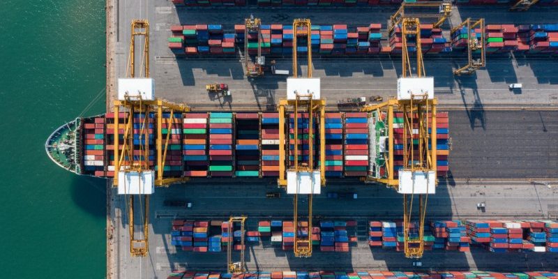 A top-down aerial view of a large container ship docked at a busy maritime terminal, being loaded and unloaded with multicolored shipping containers by tall yellow gantry cranes.